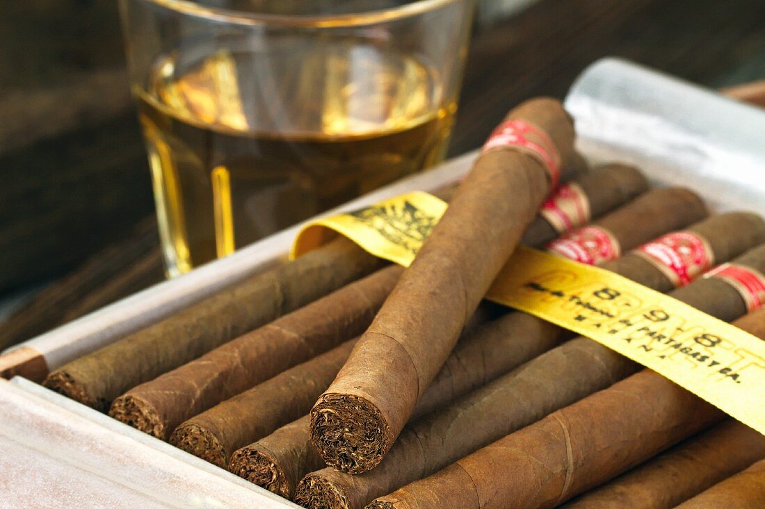 A box of Cuban cigars in front of a glass of rum