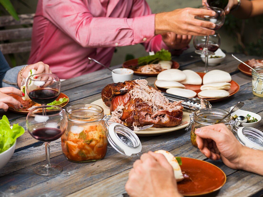 Friends enjoying roast pork with rolls, salad, kimchi and red wine at a garden table