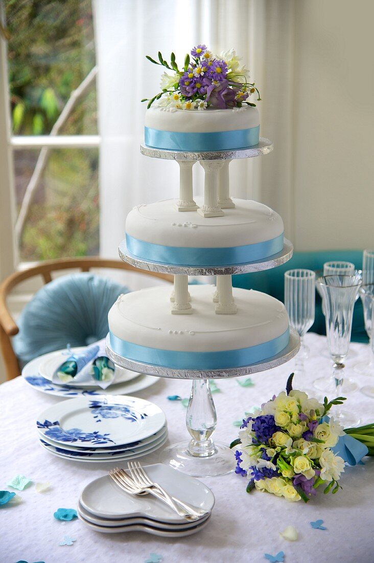 A three-tier, blue and white wedding cake decorated with flowers on a festively laid table