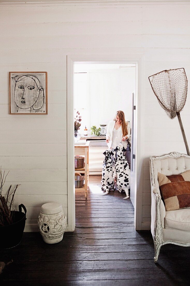 Fishing net and vintage armchair against white wooden wall, dark brown wooden floor and woman with two Dalmatians in kitchen seen through doorway