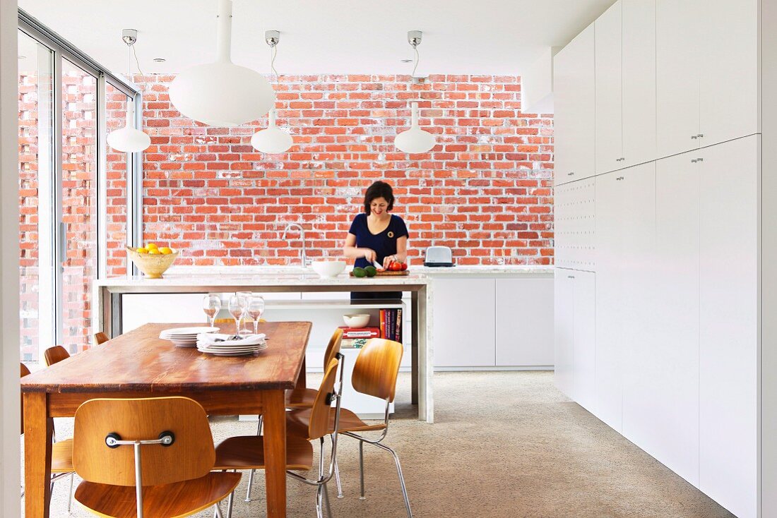Dining area in front of kitchen counter, woman preparing food, exposed brick wall, tall white fitted cupboards and classic chairs in modern, open-plan kitchen