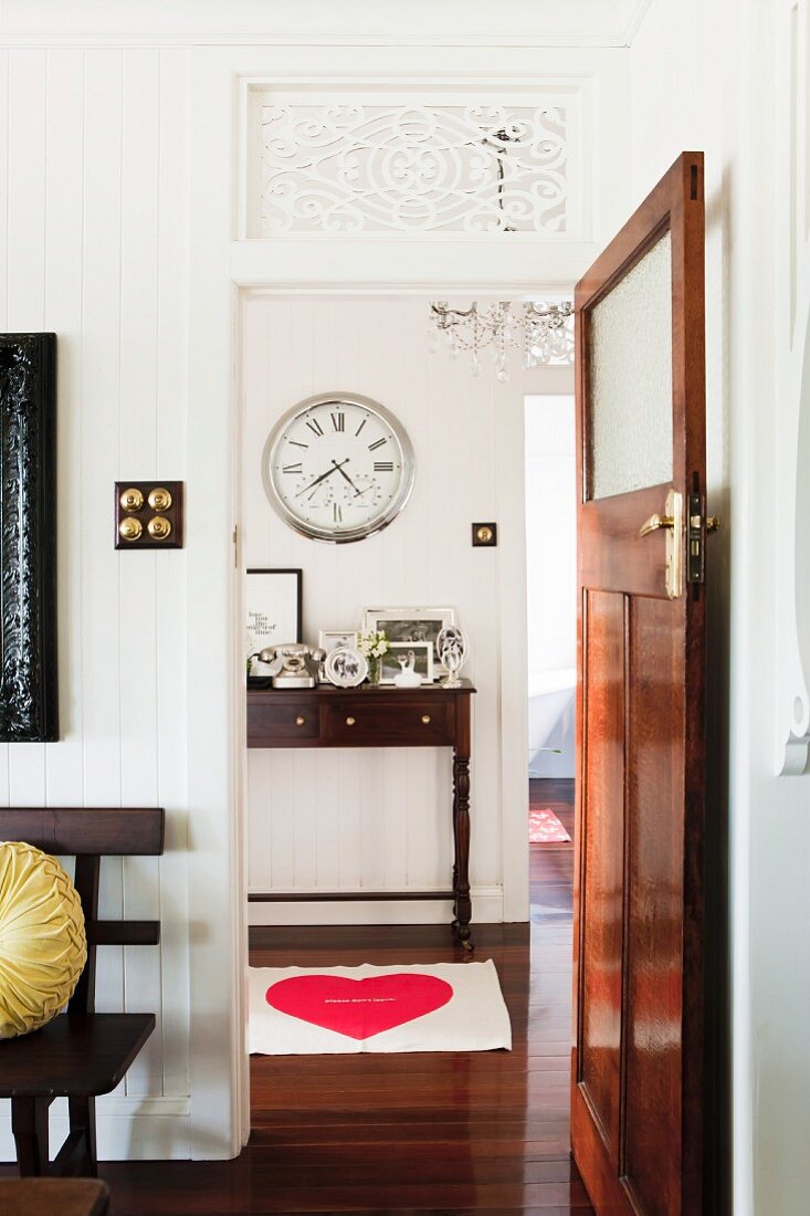 View of antique console table and wall clock with Roman numerals through open, restored interior door with transom window