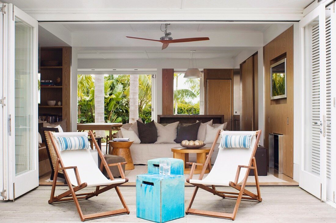 Wooden deckchairs and pale blue vintage wooden box used as side table in front of open-plan living area of bungalow with sand-coloured walls