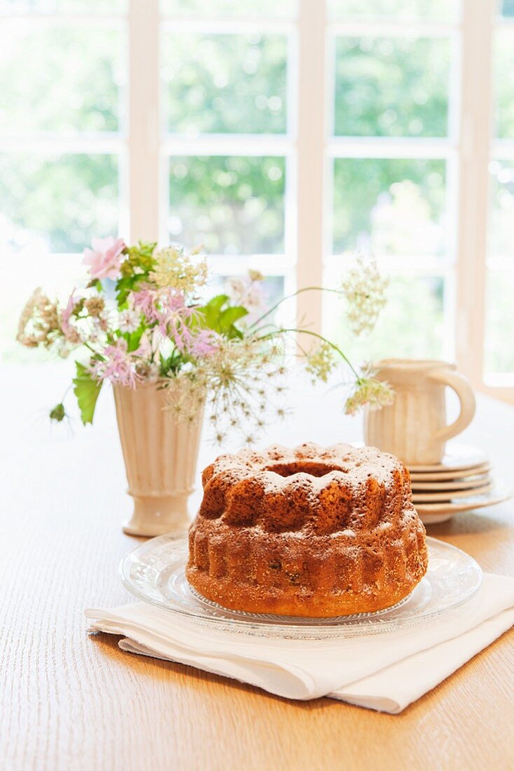 A Bundt cake with icing sugar on a table in front of a window