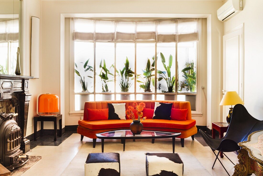 Retro-style seating area with curved orange sofa, cowhide stools, Butterfly Chair and antique fireplace to one side