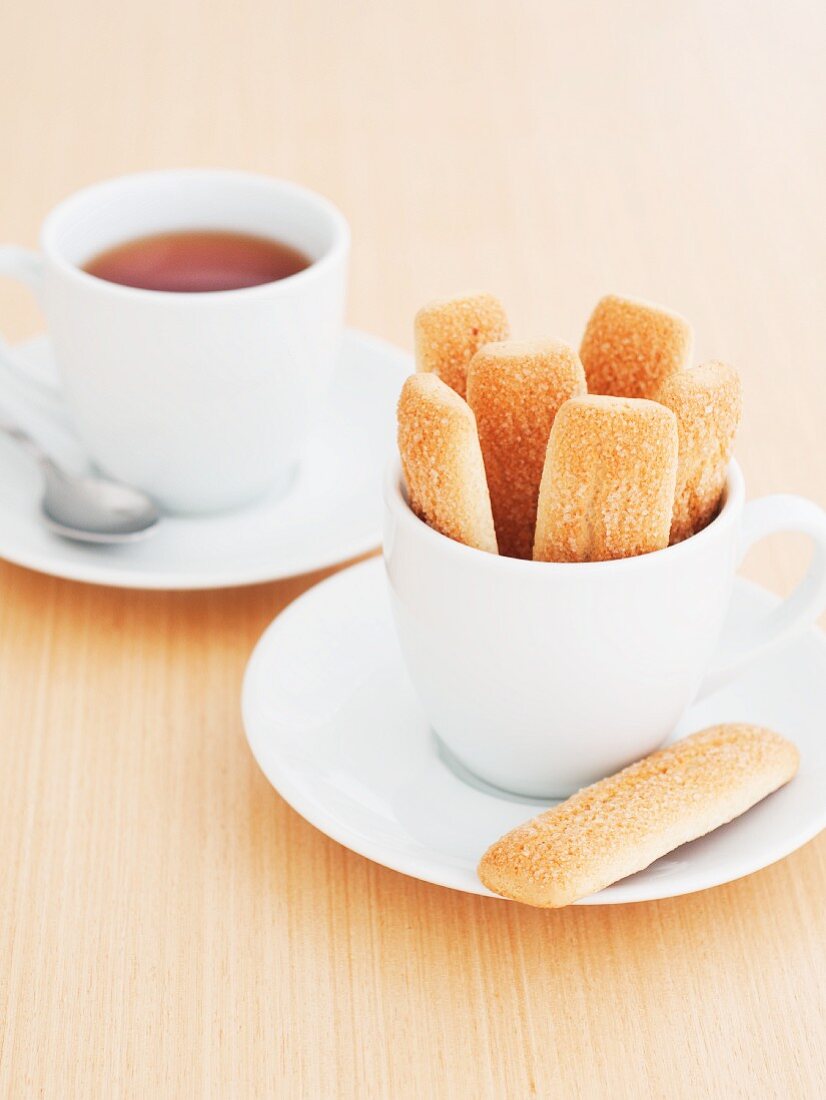 Sponge finger biscuits in a white cup and a cup of tea in the background