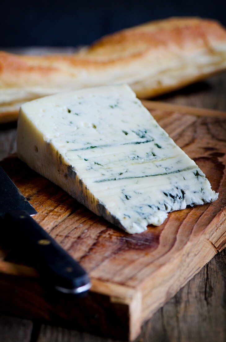 A slice of Gorgonzola on a wooden board with a baguette and a knife