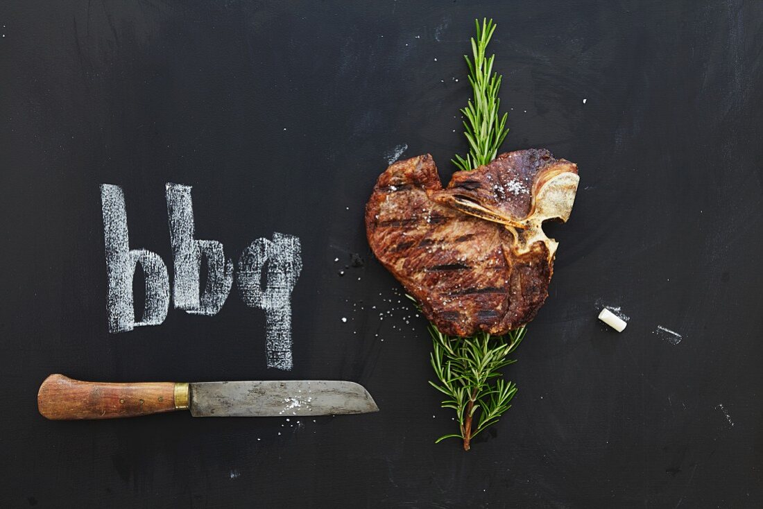 A grilled T-bone steak with rosemary and knife on a chalkboard
