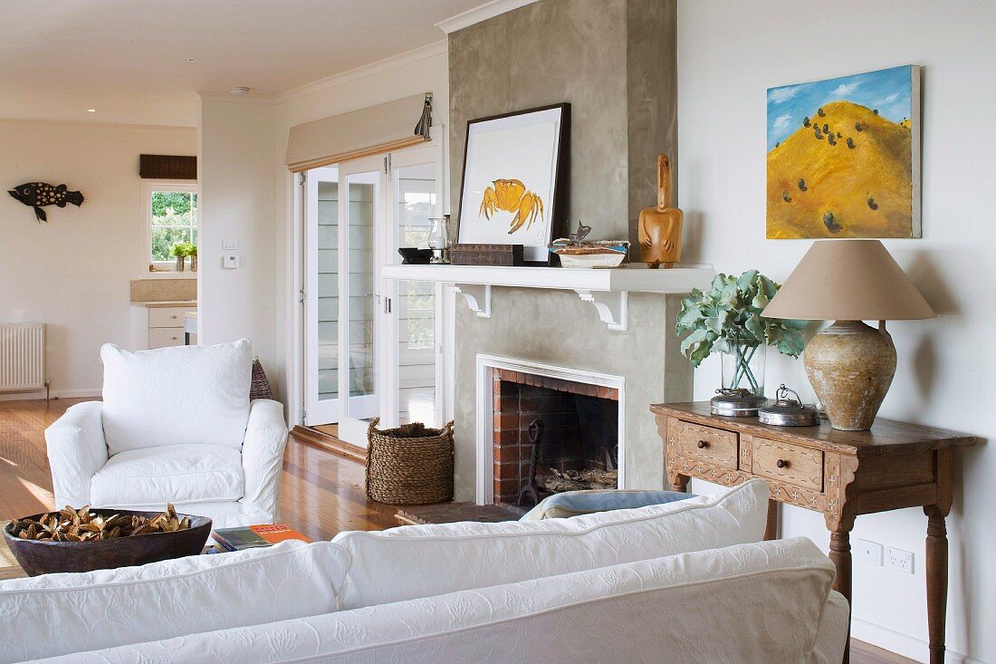 White sofa, armchair with white loose cover, open fireplace and table lamp on antique bureau in modern interior