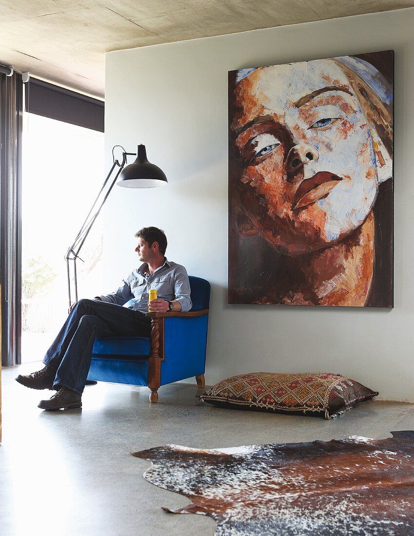 Man sitting in antique armchair upholstered in royal blue; kilim floor cushion below large portrait of woman