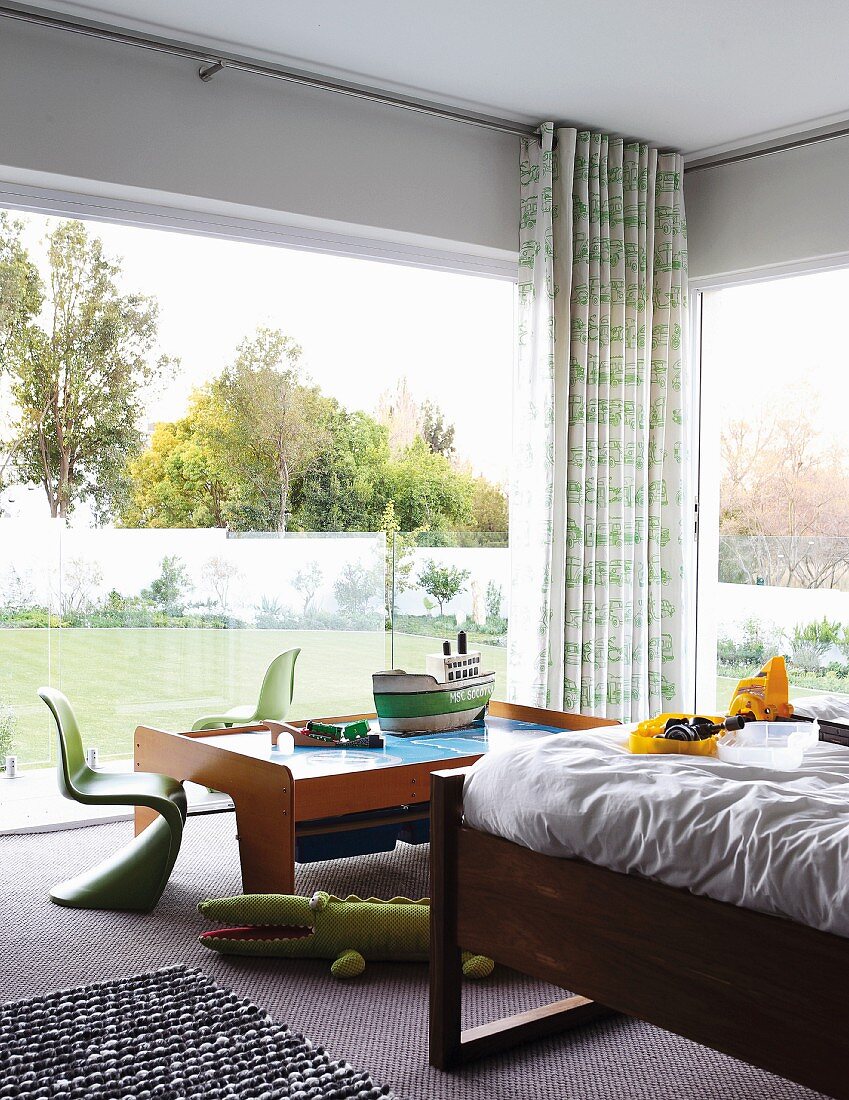 Corner of room with glass walls, partially visible bed and classic, green children's chairs around model boat on play table