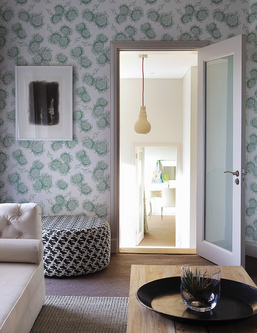 Living room with partially visible wooden coffee table, sofa, black and white patterned pouffe, floral wallpaper and view into adjacent room on lower level through open door