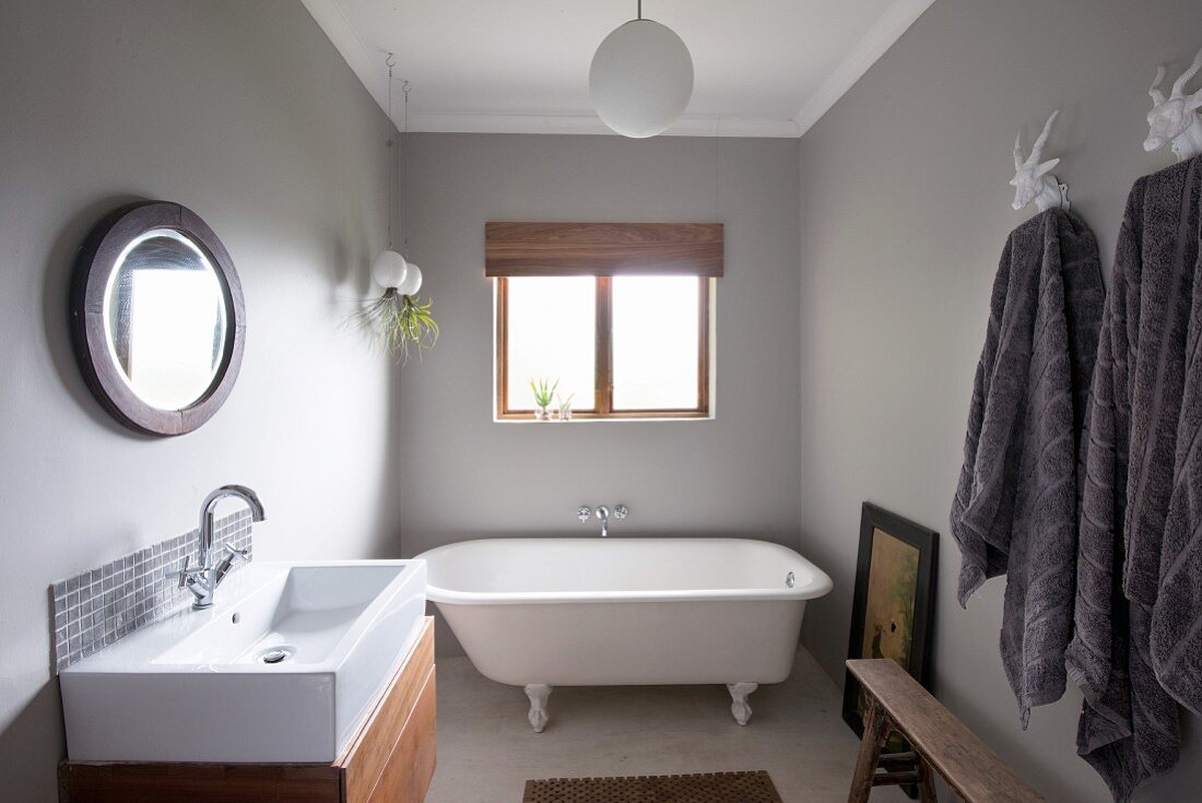 Free-standing vintage bathtub below window, modern washstand with wooden base cabinet opposite grey towels on hooks in bathroom painted pale grey