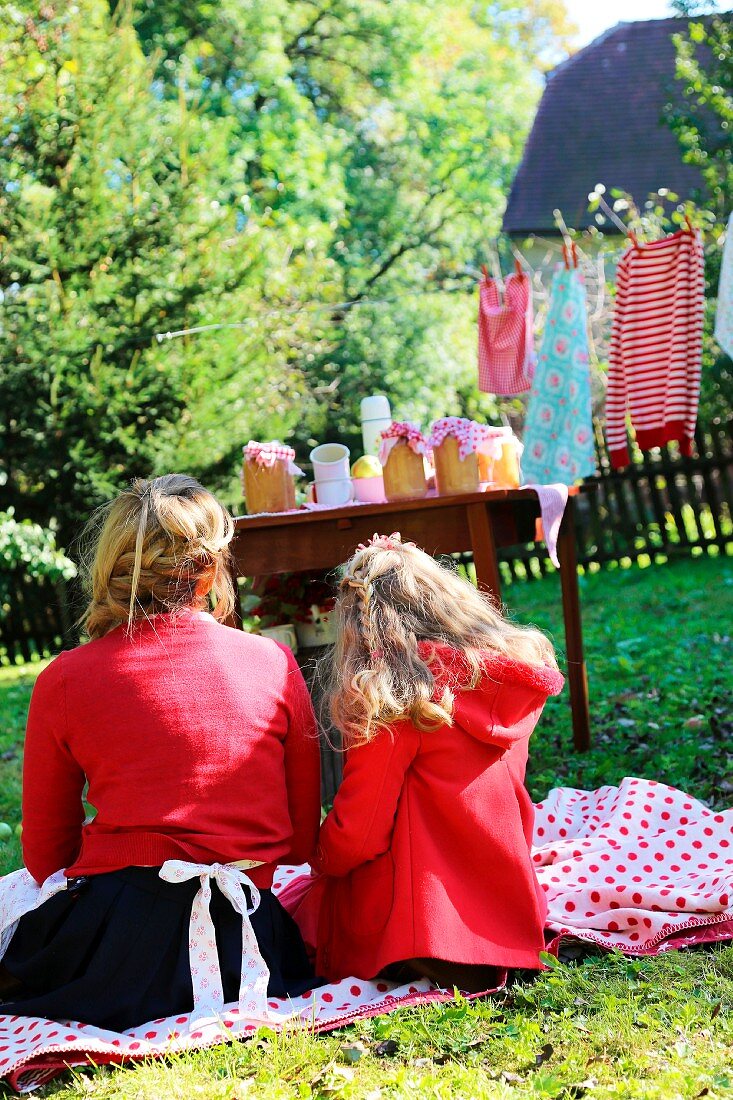 Mutter und Tochter sitzen auf Picknickdecke, im Hintergrund Tisch mit Einmachgläsern in spätsommerlicher ländlicher Stimmung