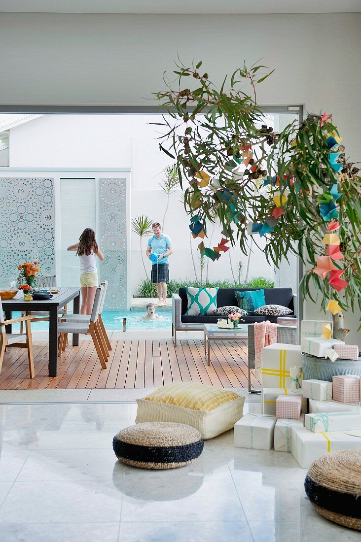 Wrapped presents below eucalyptus branch with decorated with colourful origami garlands used as Australian Christmas tree; view of family on wooden terrace next to pool