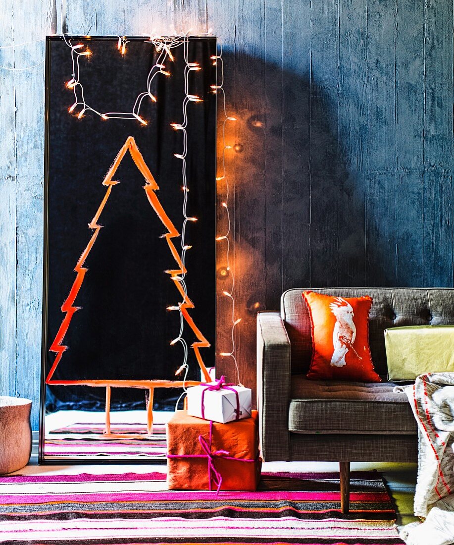 Stylised Christmas tree painted in orange paint on full-length mirror and fairy lights against blue-grey wall, colourful stripes rug and wrapped presents next to retro sofa
