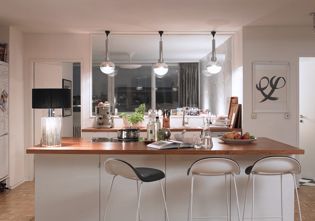 Bar stools at white counter with wooden worksurface; table lamp with black lampshade to one side and pendant lamps with spherical lampshades above kitchen counter