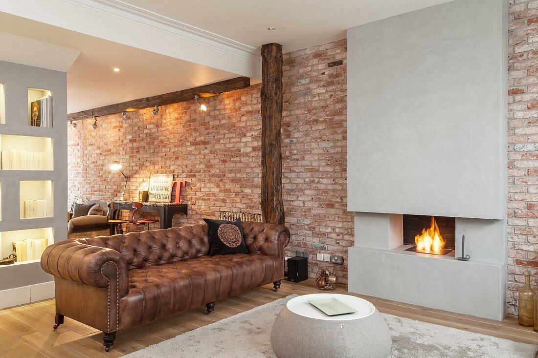 Open-plan interior with old leather sofa next to open fire; concrete chimney breast flanked by brick walls