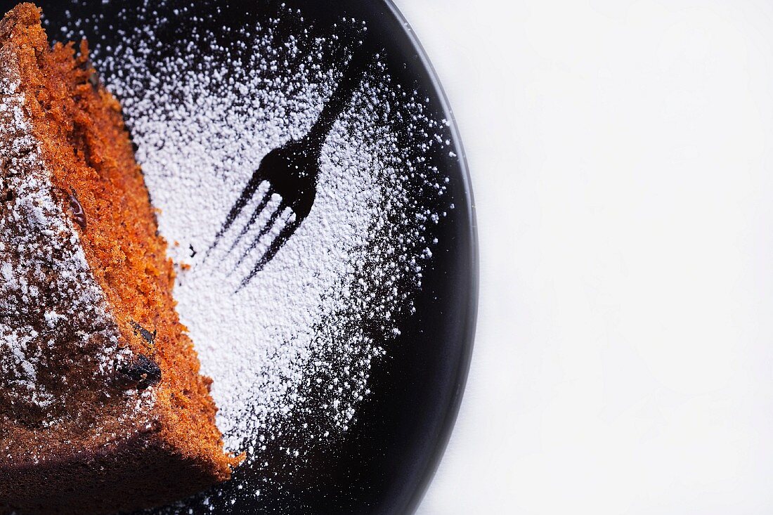 A slice of chocolate cake on a plate dusted with icing sugar with a print from a fork in the icing sugar