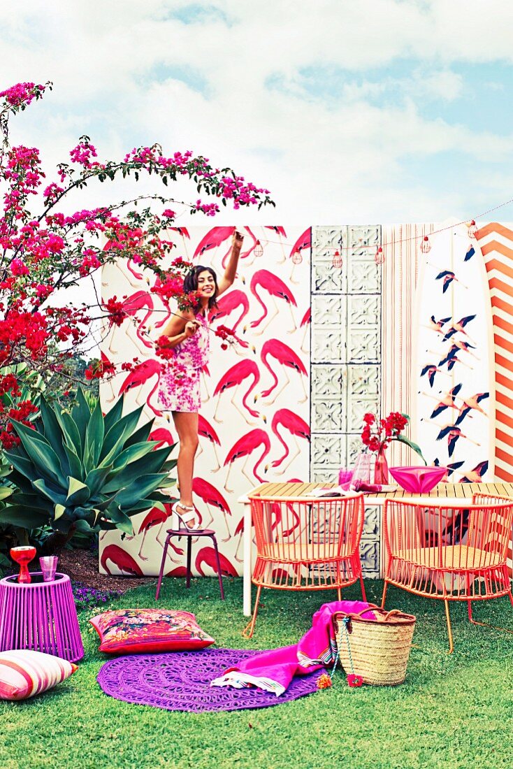 Different flamingo print patterns, outdoor furniture and accessories in shades of red and purple; woman standing on stool hanging up fairy lights