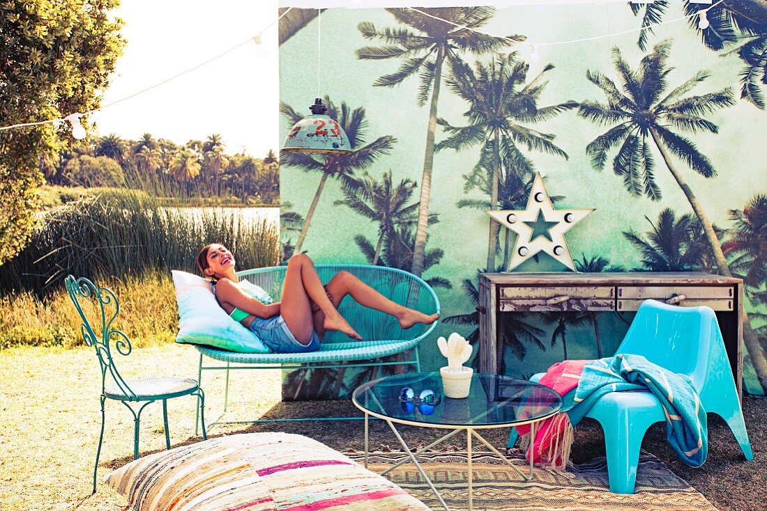 Decoration for outdoor party with combination of azure blue seating, rug and floor cushions in front of palm tree pattern on screen; young woman posing on bench