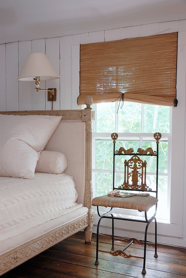 Delicate, ornate metal chair and antique bed with tall headboard next to window with half-closed, bamboo blinds