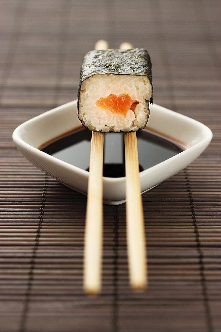 Maki sushi with salmon balanced on chopsticks over a bowl of soy sauce