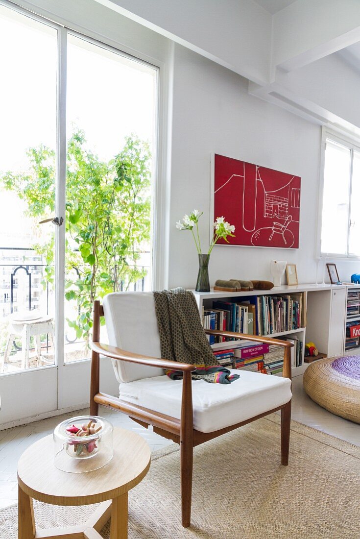 60s-style armchair with wooden frame and wooden side table in loft-style interior front of French windows with view of small potted tree