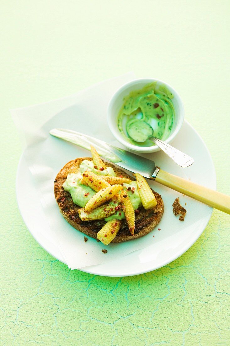 Slice of grilled bread topped with avocado cream and baby corn cobs