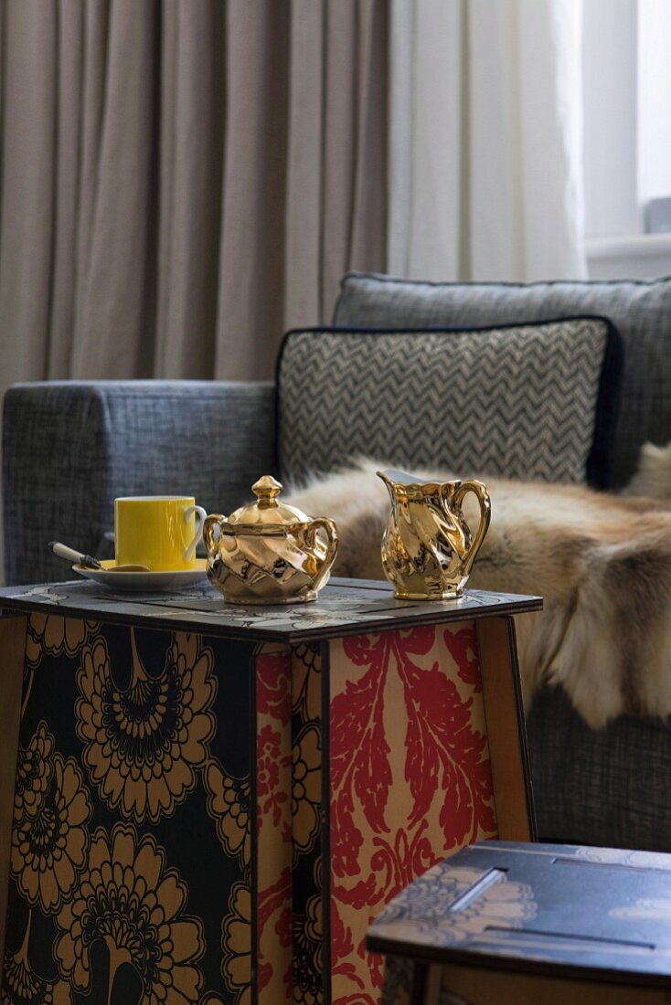 Gilt porcelain milk jug and sugar bowl on side table made from interlocking panels with floral, Art-Nouveau patterns in front of fur blanket on sofa