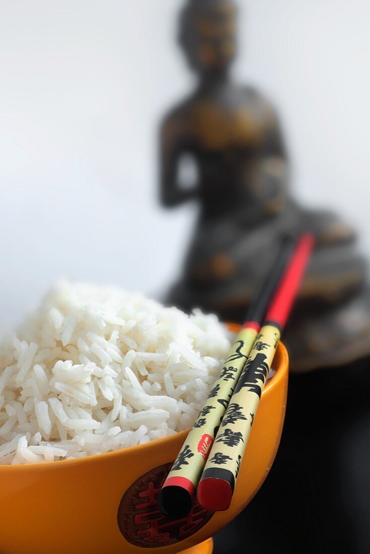 A bowl of rice and chopsticks in front of a statue of Buddha