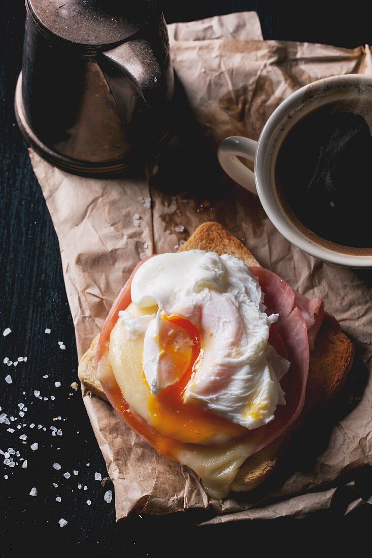 Aufsicht auf Schinken-Käse-Toast mit pochiertem Ei auf Pergamentpapier, dazu Kaffee