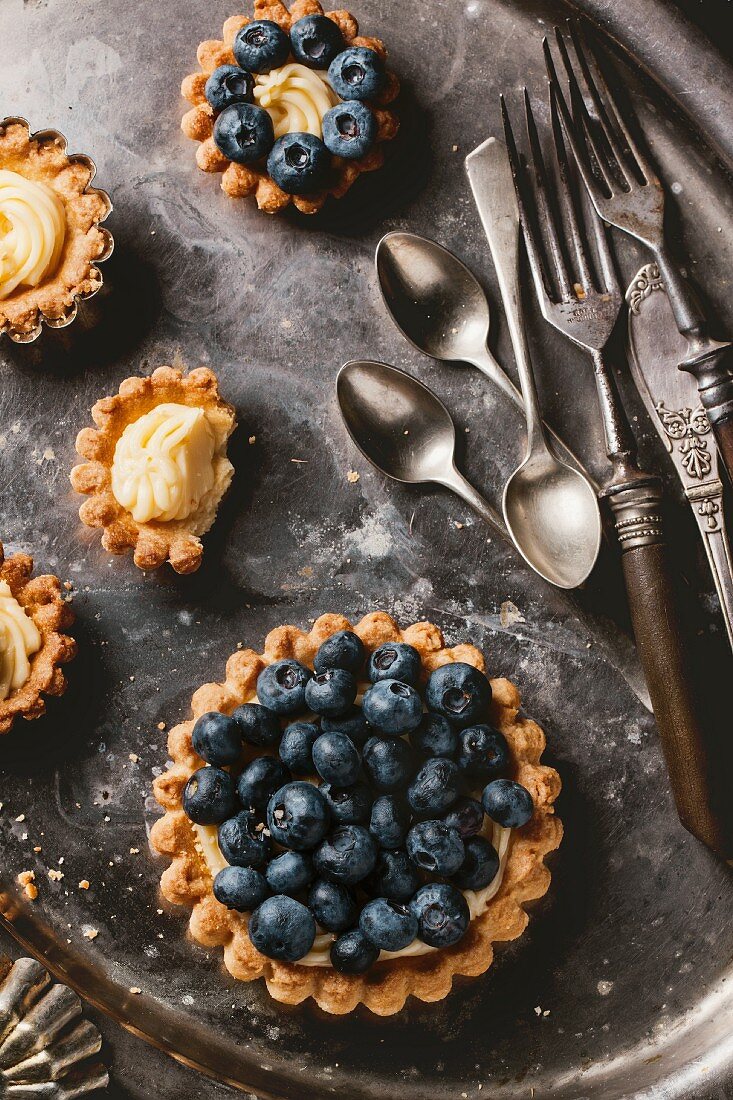 Silver cutlery and various different sized blueberry tartlets with lemon cream, one with a bite taken out