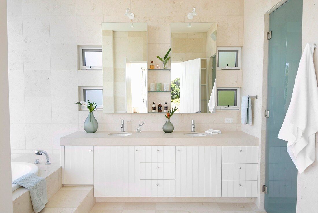 Washstand with stone top, twin countertop basins and white cabinets below mirrored cabinet in bright, modern bathroom