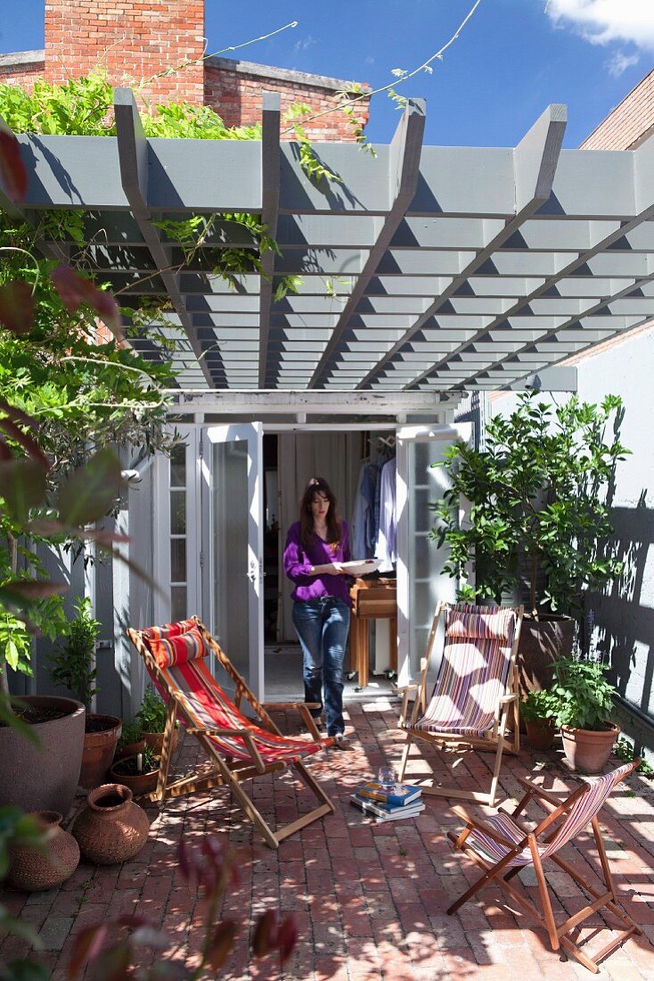 Liegestühle auf Terrasse, unter Pergola aus grau lackierter Kassetten-Holzkonstruktion, im Hintergrund Frau vor Wohnhaus