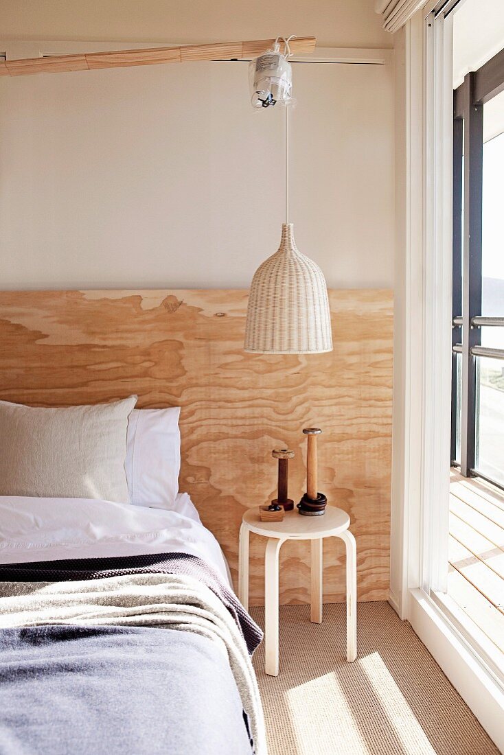 Pendant lamp with wicker lampshade above side table next to bed against wall with rustic wooden panel in sunny bedroom