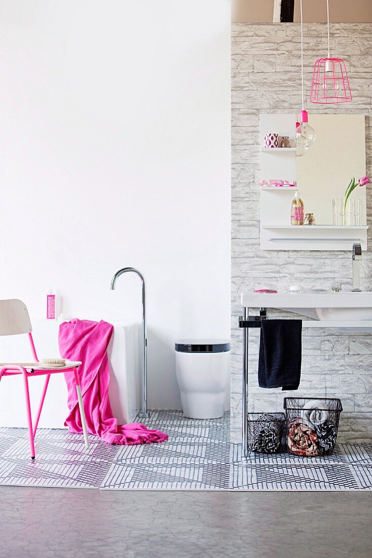 Baskets below minimalist washstand, chair with pink metal frame and bright pink towel on bathtub with floor-mounted tap fittings in bathroom