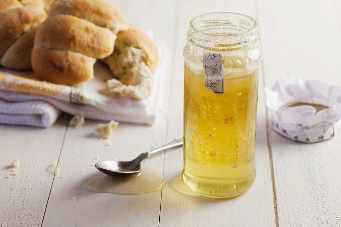 Honig mit Honigwabe im Glas, daneben Brötchen auf Geschirrtuch