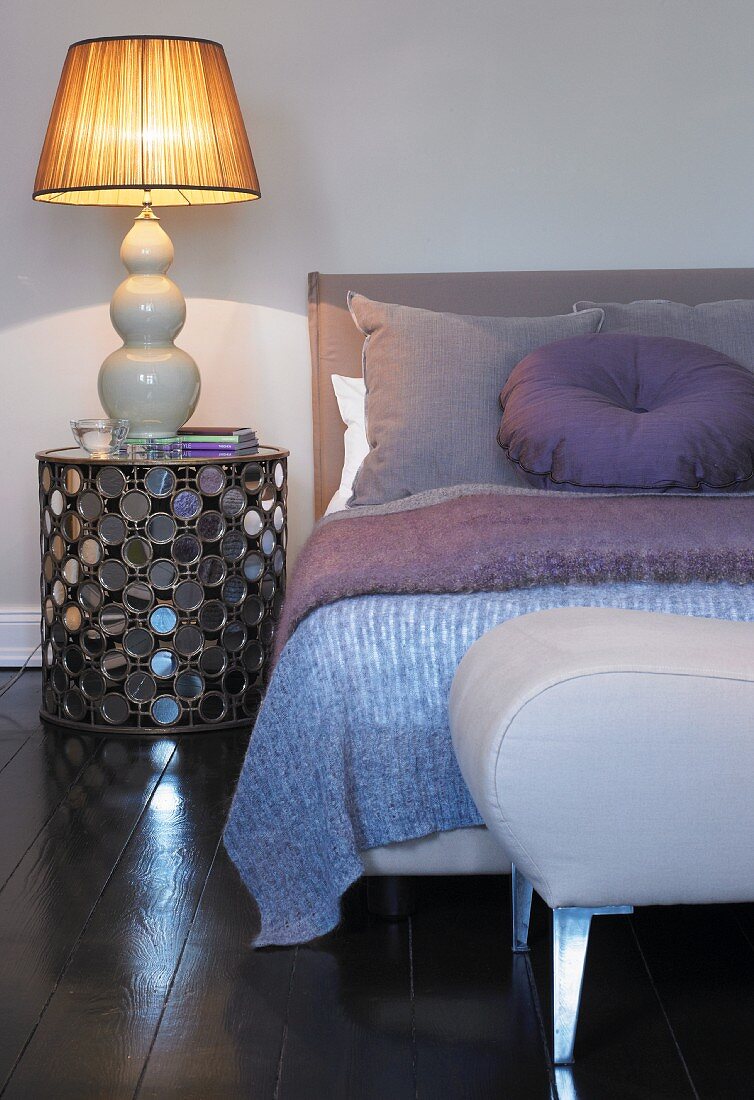 Lilac cushion and bedspread on bed, lamp on bedside cabinet and stool at foot of bed in bedroom
