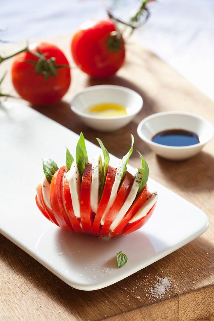 Tomate fächerförmig geschnitten, mit Mozzarella und Basilikum gefüllt, dahinter Balsamico und Olivenöl