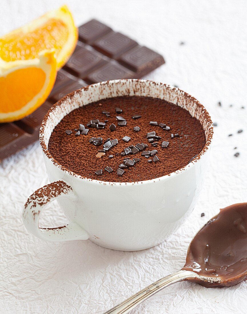 Schoko-Orangen-Pudding in Tasse, bestäubt mit Kakaopulver, dahinter Zutaten