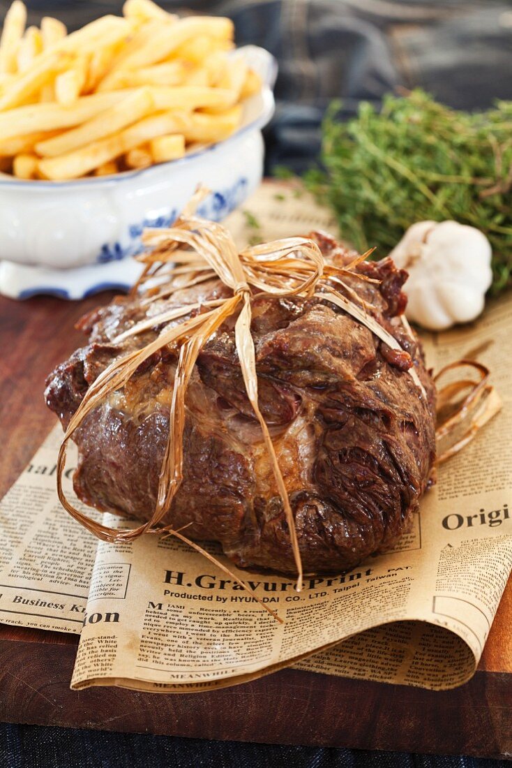 A slice of roast beef tied with kitchen twine on a piece of newspaper with a bowl of chips in the background