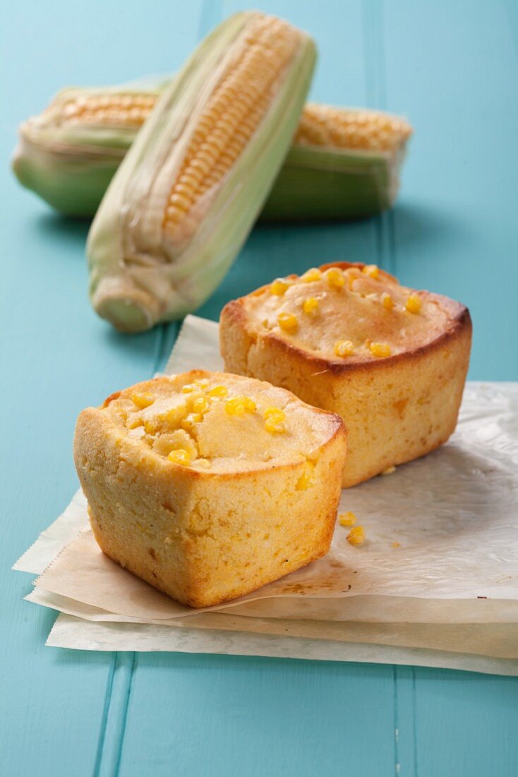 Two small, square corn cakes on a piece of paper with corn cobs in the background
