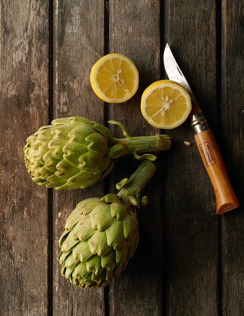 Two artichokes, a halved lemon and a knife on a wooden surface