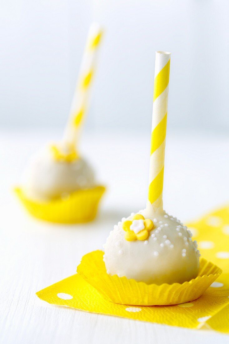 weiße Cake Pops mit gelb gestreiftem Stiel in gelben Papierförmchen auf gepunkteter Serviette