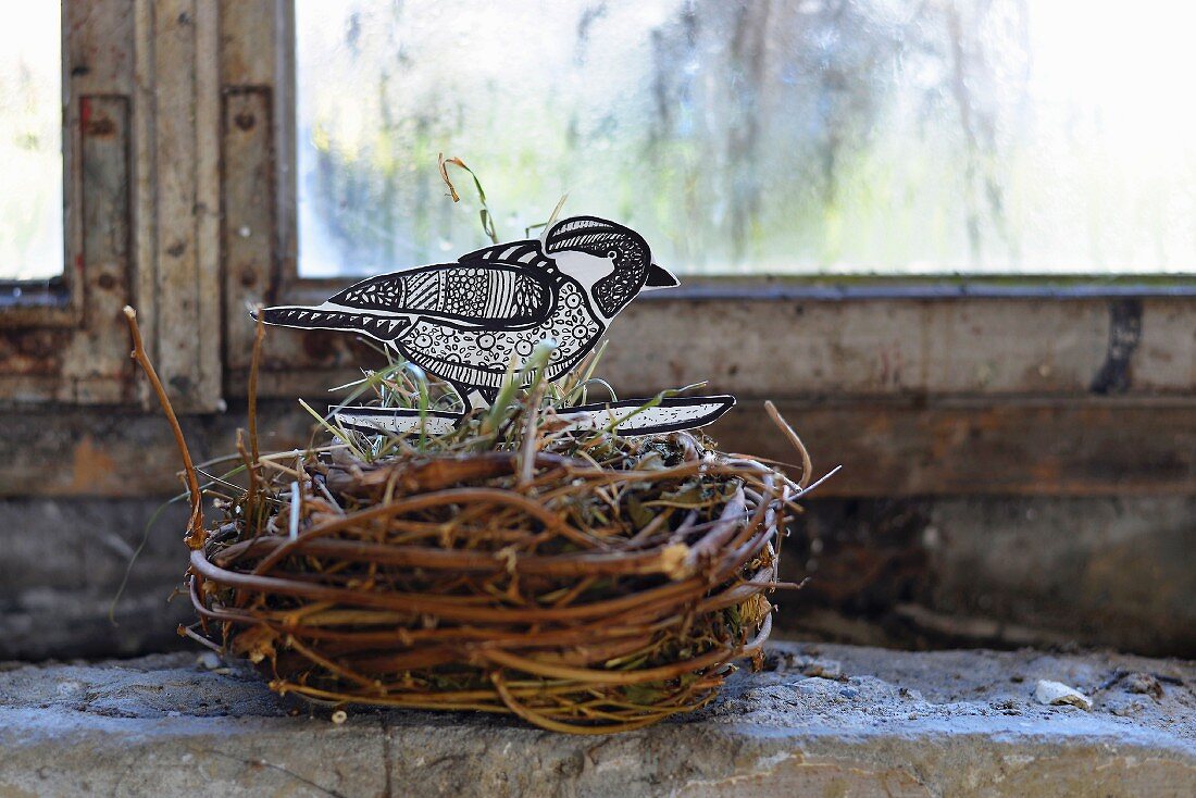 Vogelnest aus Weidenzweigen mit selbstgebasteltem Vogel aus Papier auf Ablage am Fenster