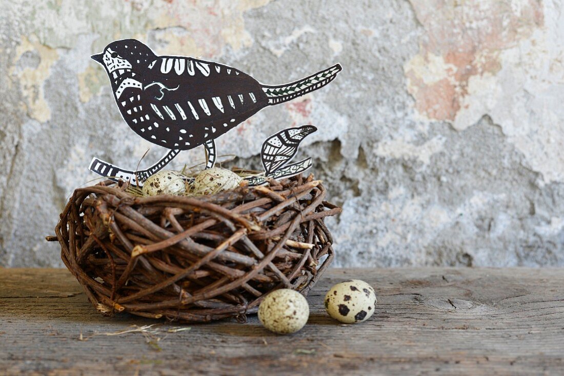 Hand-crafted paper bird and quail eggs in birds' nest on wooden surface against wall with peeling paint