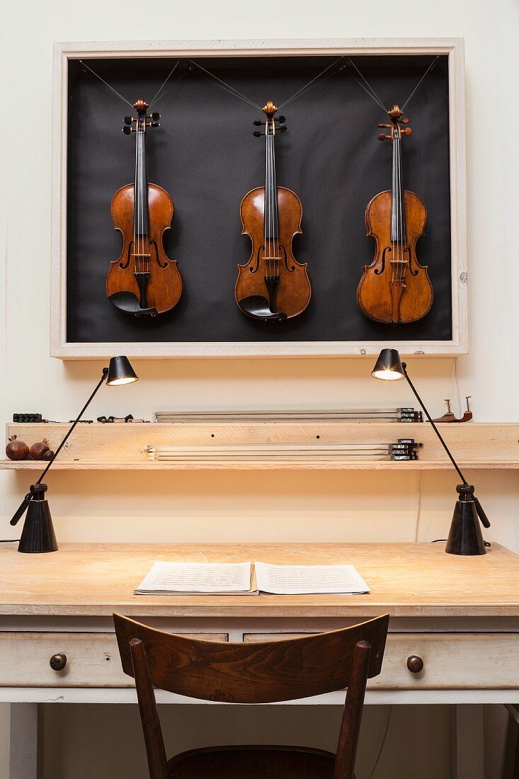 Three violins in display case above vintage table with drawers lit by tow modern desk lamps