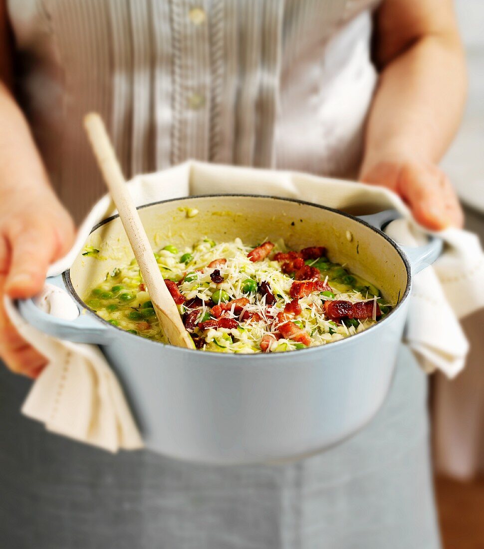 Frau hält Topf mit Risotto mit Zucchini, Erbsen und Speck