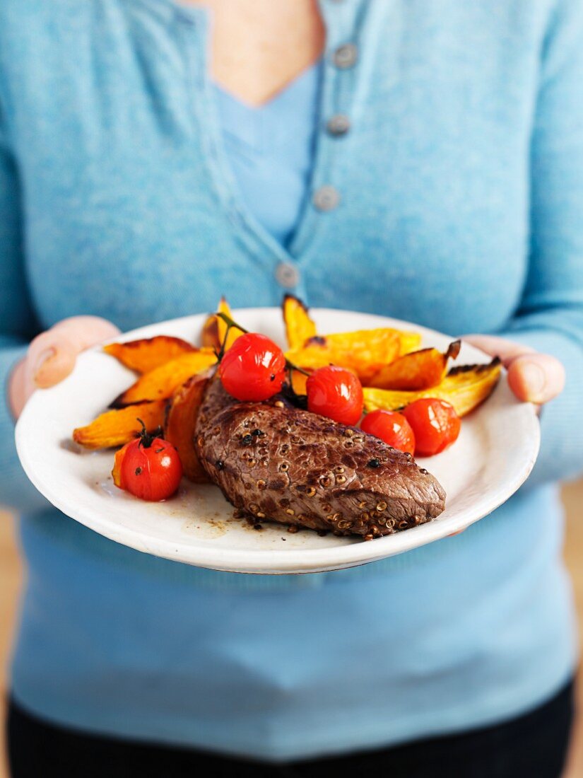 A woman holding a plate of steak, tomatoes and sweet potato wedges
